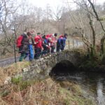 group of pupils in outdoor clothing standing over a small arched stone bridge and looking into the water